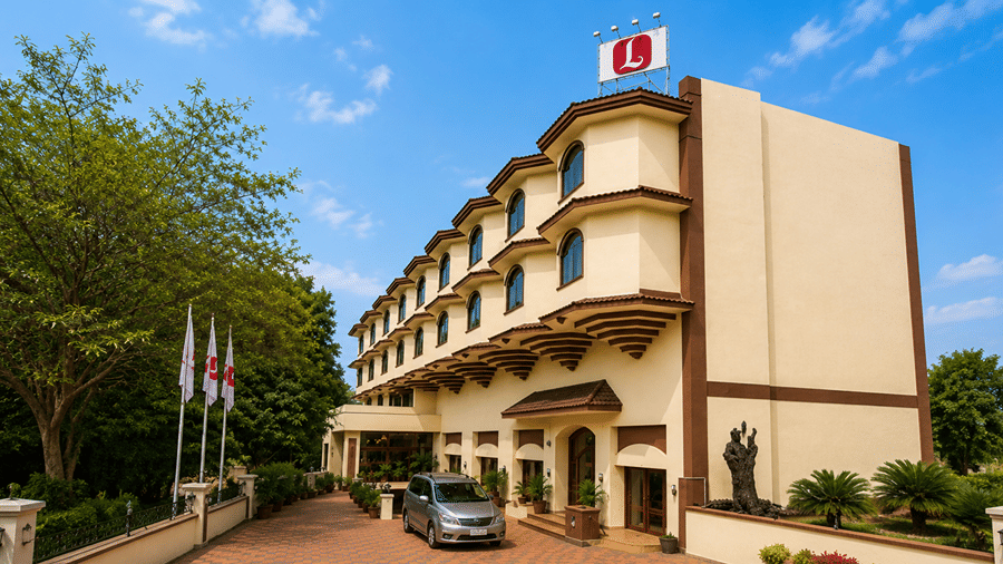 Exterior view of Lords Inn Vadodara showing a cream-colored multi-story building with arched window details, a rooftop logo, a parked car in the entrance driveway, and surrounding green trees under a bright blue sky.
