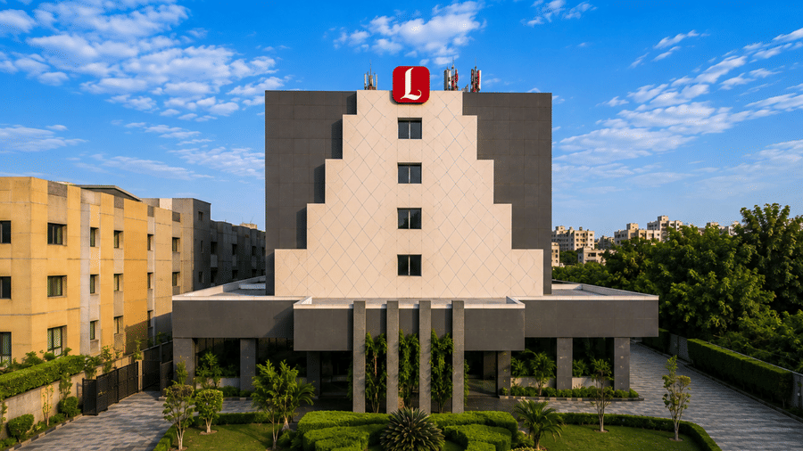 Exterior daytime view of Lords Plaza Ankleshwar featuring a stepped multi-story architectural facade, red rooftop brand logo, landscaped entrance, and a bright blue sky.