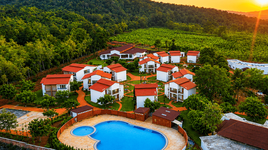 Aerial view of Lords Sahyadri Resort, Sagara - Shivamogga featuring red-roofed cottages nestled among dense green forestry, with a large blue swimming pool and outdoor sports court in the foreground.