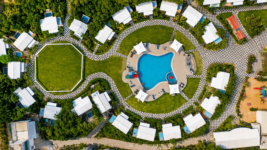 Aerial top-down view of Lords SKD Resort, Salangpur featuring white cottages arranged around a central swimming pool and a circular green lawn surrounded by lush trees.