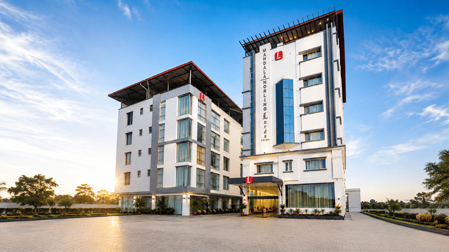 Exterior view of Mandala Norling Lords Prime Kathmandu featuring two modern white multi-story hotel wings with blue glass details, pitched roofs, and a wide paved forecourt under a bright blue sky.