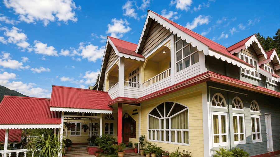 Exterior view of Pemaling Lords Inn Dirang featuring sloped red roofs, yellow and grey facade, and white trim against a backdrop of green hills and blue sky.
