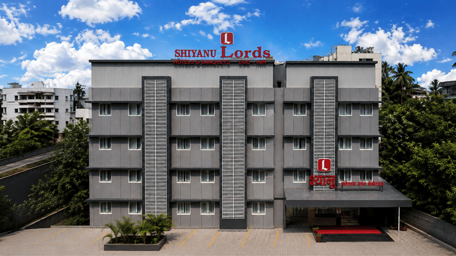 Exterior view of Shyanu Lords Eco Inn Airoli featuring a multi-story gray hotel building with rooftop signage, a ground-level entrance, and surrounding greenery under a blue sky.