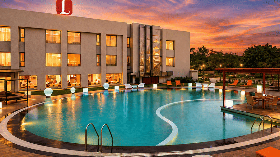 Exterior sunset view of Top3 Lords Resort Bhavnagar featuring a large illuminated outdoor swimming pool, poolside lounge chairs, wooden pergola, and resort building facade under a vibrant twilight sky.