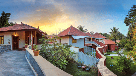 Several cottages at Hoysala Village Resort, Hassan with red-tiled roofs sit amongst trees under a glowing sunset sky, bordered by a long, winding wall.