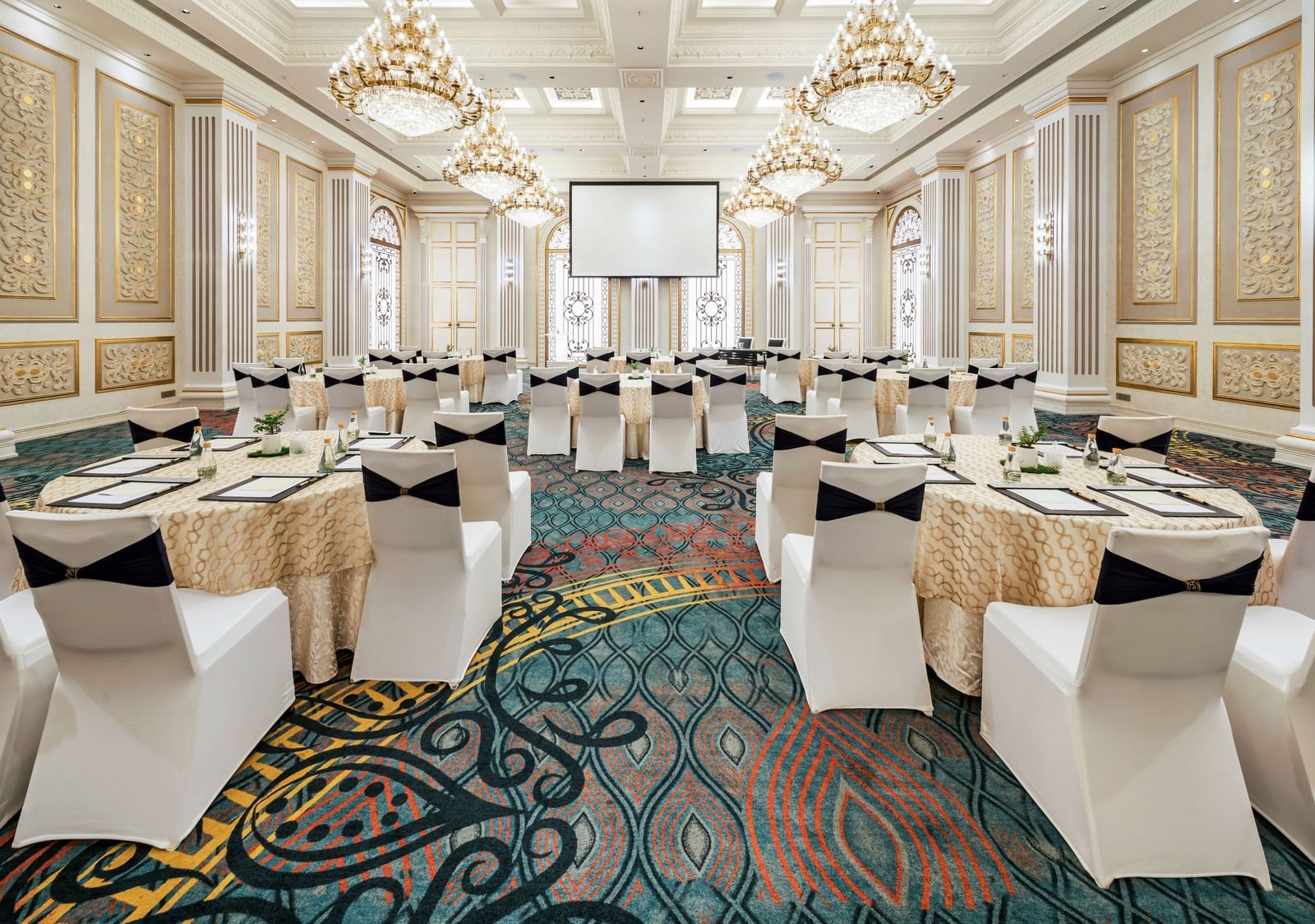 An expansive event hall at Grand Chennai at GRT Hotels featuring a striking patterned carpet, filled with rows of white chairs facing a central point, perfect for a large-scale presentation or formal gathering.