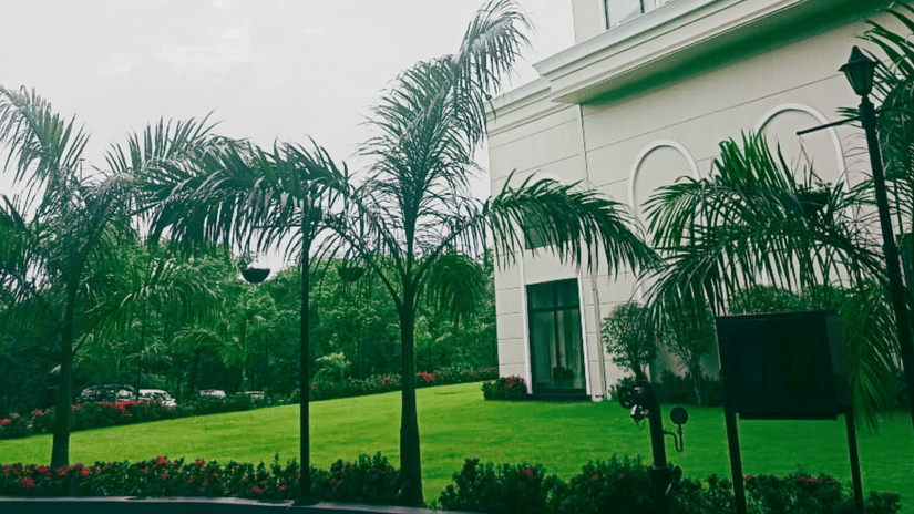 Landscaped garden at Polo Hotel, Agartala with row of foxtail palm trees, red flowerbeds, manicured lawn and arched white facade.