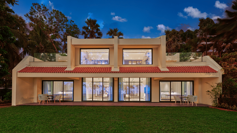 An exterior view of the villa at The Resort, Mumbai, with a manicured garden in front and trees surrounding it.