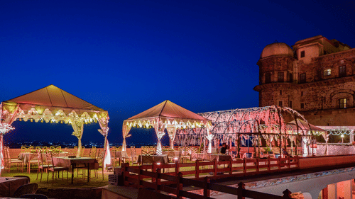 2 tented structures and a larger illuminated structure stand on a terrace next to a building at Tijara Fort-Palace - 19th Century, Alwar.