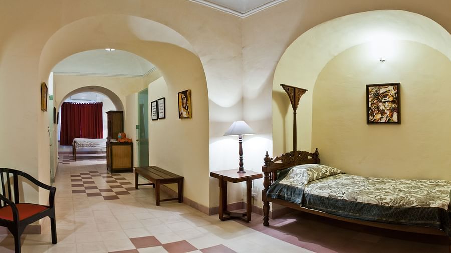 Hotel hallway with arched doorways, a single bed with a lamp, a chair, and a bench at Tijara Fort-Palace, Alwar.