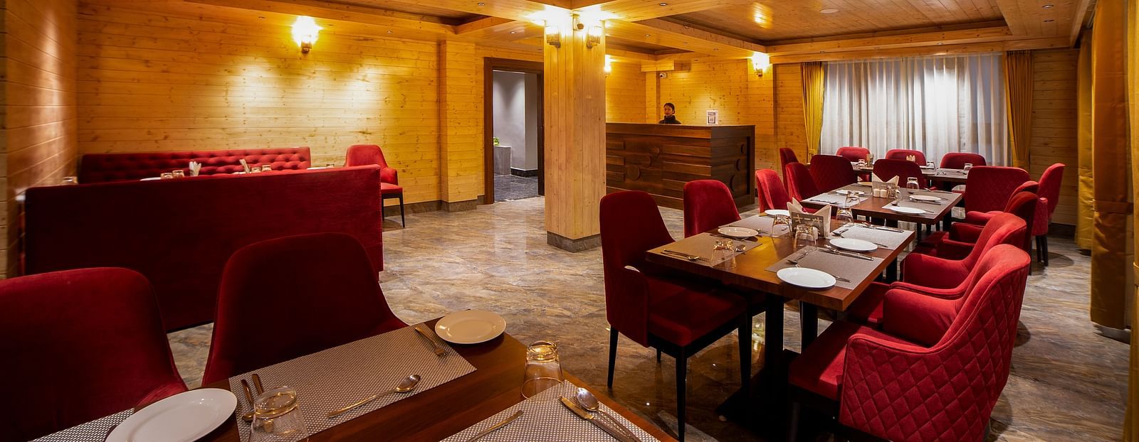 Elegant restaurant dining area with warm wooden walls and ceiling, featuring red upholstered chairs, dark wooden tables set with white placemats and cutlery, and soft ambient lighting from ceiling-mounted fixtures - Yashshree Lachen.
