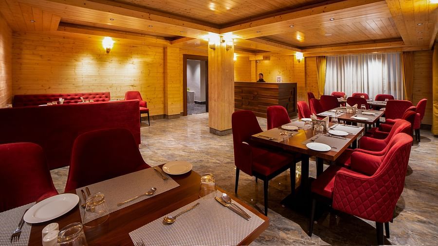 Elegant restaurant dining area with warm wooden walls and ceiling, featuring red upholstered chairs, dark wooden tables set with white placemats and cutlery, and soft ambient lighting from ceiling-mounted fixtures - Yashshree Lachen.