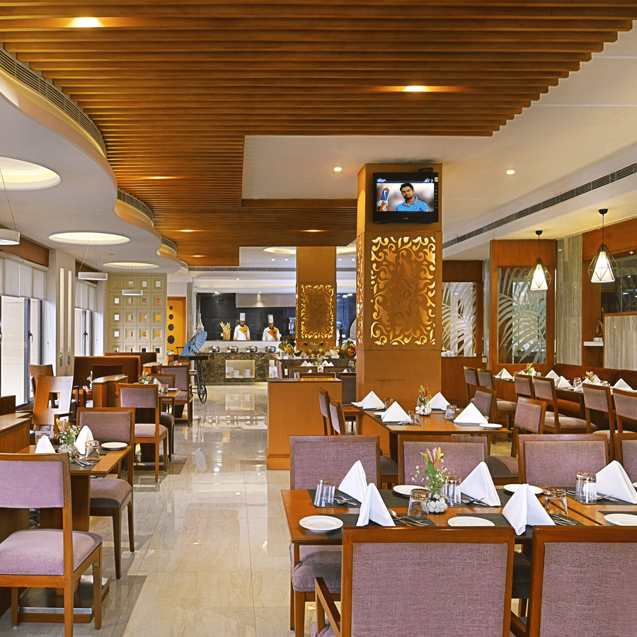 Coffee Shop restaurant at The Bristol Hotel, Gurgaon with tables set, mauve upholstered chairs, a wood-slat ceiling, pendant lights, and a buffet counter.