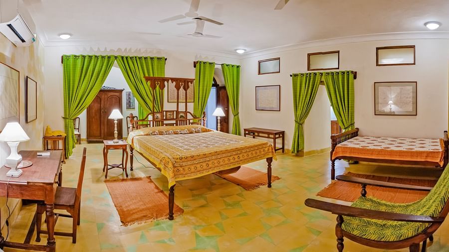 A spacious bedroom with two beds featuring patterned bedspreads, wooden furniture including wardrobes and bedside tables with lamps, a desk with a chair, a lounge chair, windows with curtains, and framed pictures on the walls at Tijara Fort-Palace - 19th C