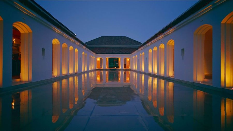 Tranquil courtyard at dusk with glowing yellow lights illuminating white arches, reflected in a long central water feature leading towards a building with a dark tiled roof - Marasa Sarovar Premiere, Bodhgaya