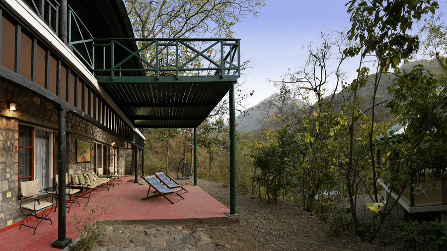 Stone architecture of the main building with a long red porch area and green wooden railings overlooking the forest canopy at Neemrana’s Glasshouse on The Ganges offering a serene spot to sit and admire the natural beauty of the river valley