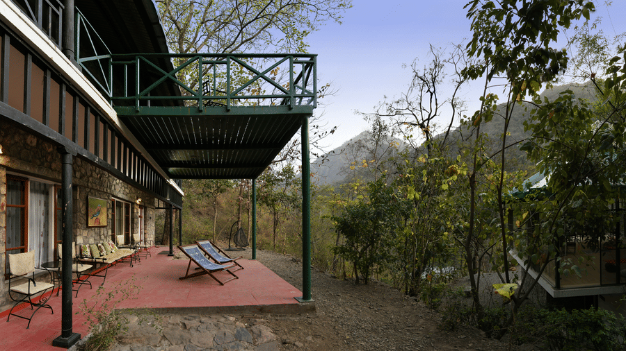 Stone architecture of the main building with a long red porch area and green wooden railings overlooking the forest canopy at Neemrana’s Glasshouse on The Ganges offering a serene spot to sit and admire the natural beauty of the river valley