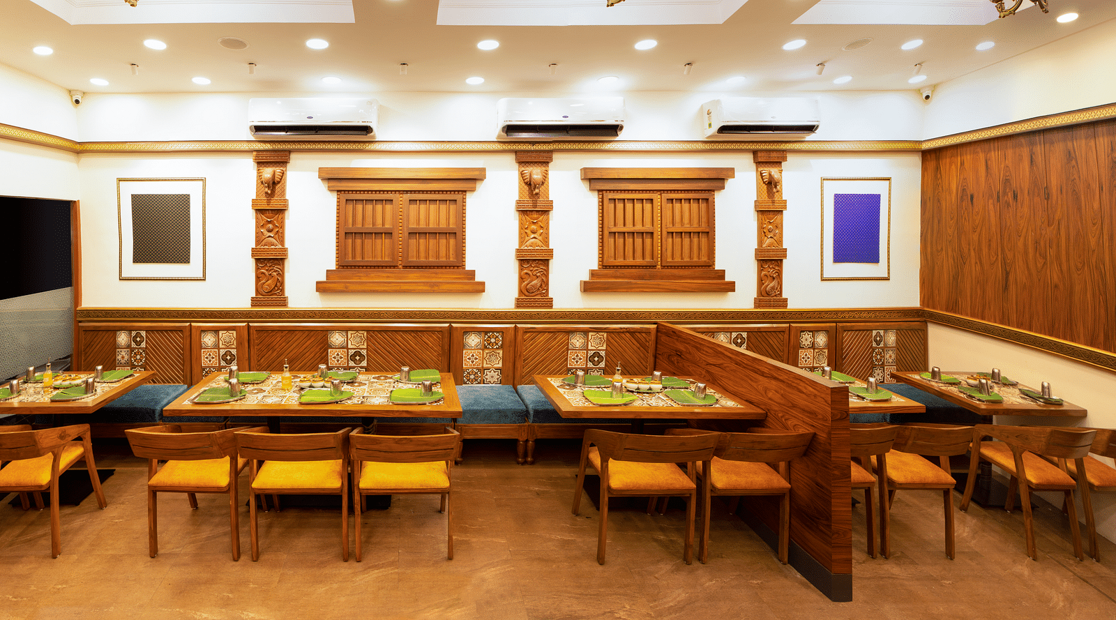 A dining hall with wooden tables set with plates and glasses under elegant lighting | Kamats India
