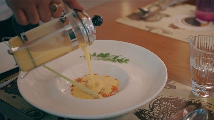 A person pours creamy yellow soup from a French press into a white bowl garnished with herbs, vegetables, and a lemongrass stalk, placed on a wooden dining table - Marasa Sarovar Premiere, Bodhgaya