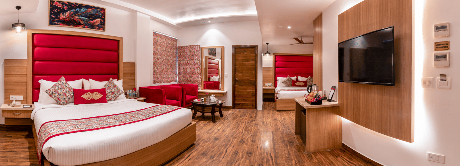 Side view of a Family Room at Lords Inn Amritsar with a king bed, red headboard, wall-mounted TV, wooden flooring, and warm ambient lighting.