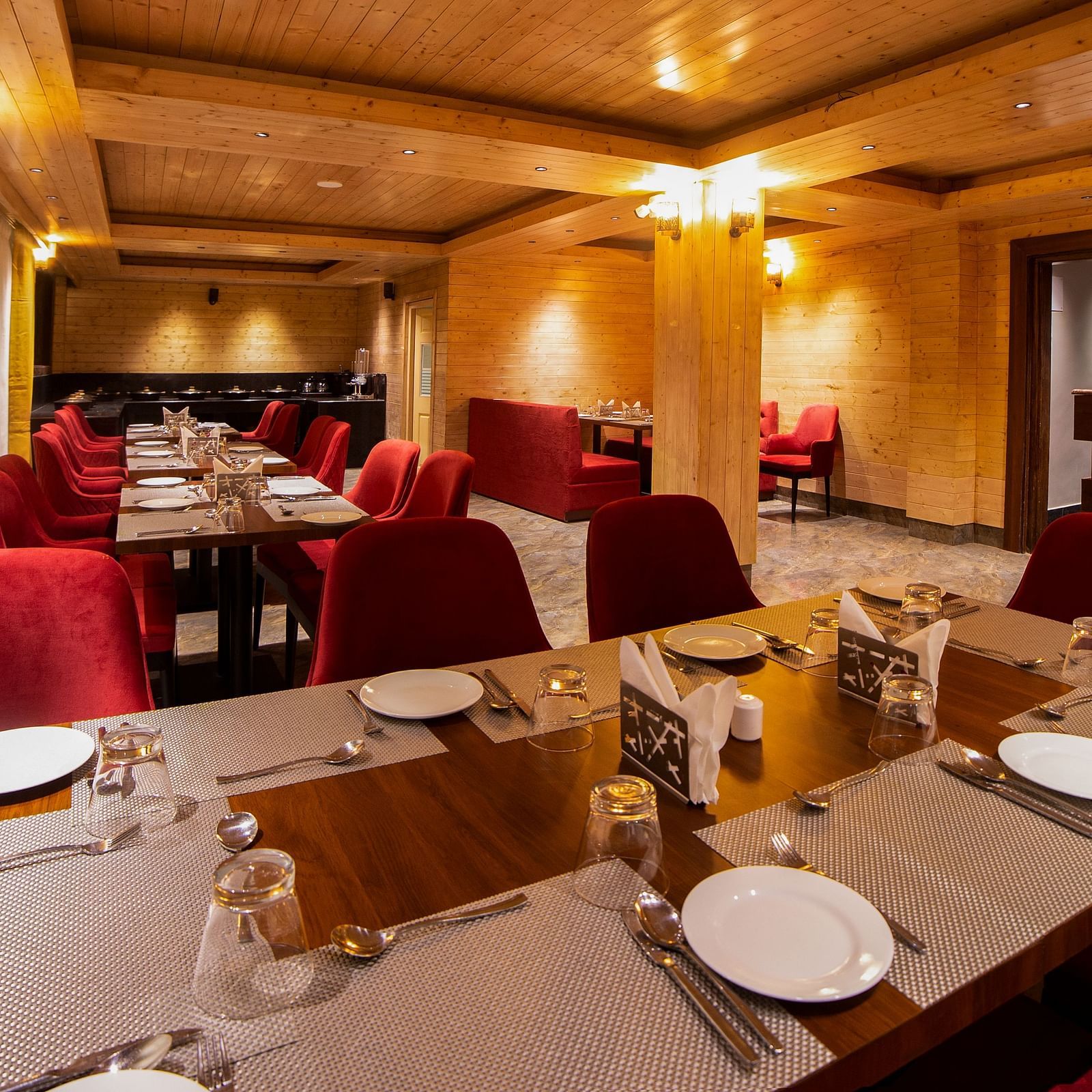 Elegant restaurant interior featuring neatly set wooden tables with white placemats and red upholstered chairs, warm lighting, wooden ceiling panels, and curtained windows lining one side of the room - Yashshree Lachen.