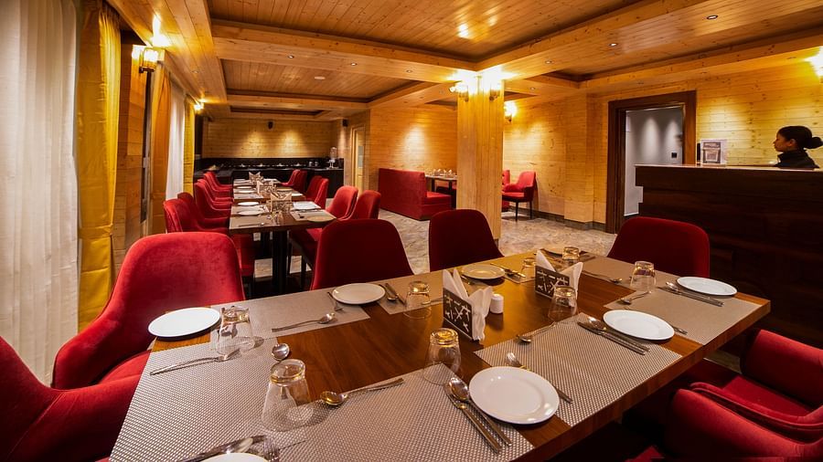 Elegant restaurant interior featuring neatly set wooden tables with white placemats and red upholstered chairs, warm lighting, wooden ceiling panels, and curtained windows lining one side of the room - Yashshree Lachen.