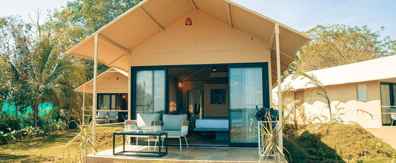 Front view of a canvas tent with patio seating, surrounded by dry grass and trees at Fireflies Resorts, Kabini.