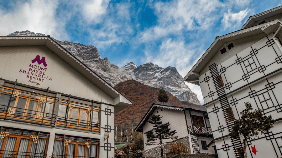 Mount Bayul Shangri-La Resort in Lachung features traditional-style buildings with wooden balconies, set against a mountainous backdrop beneath a partly cloudy sky.