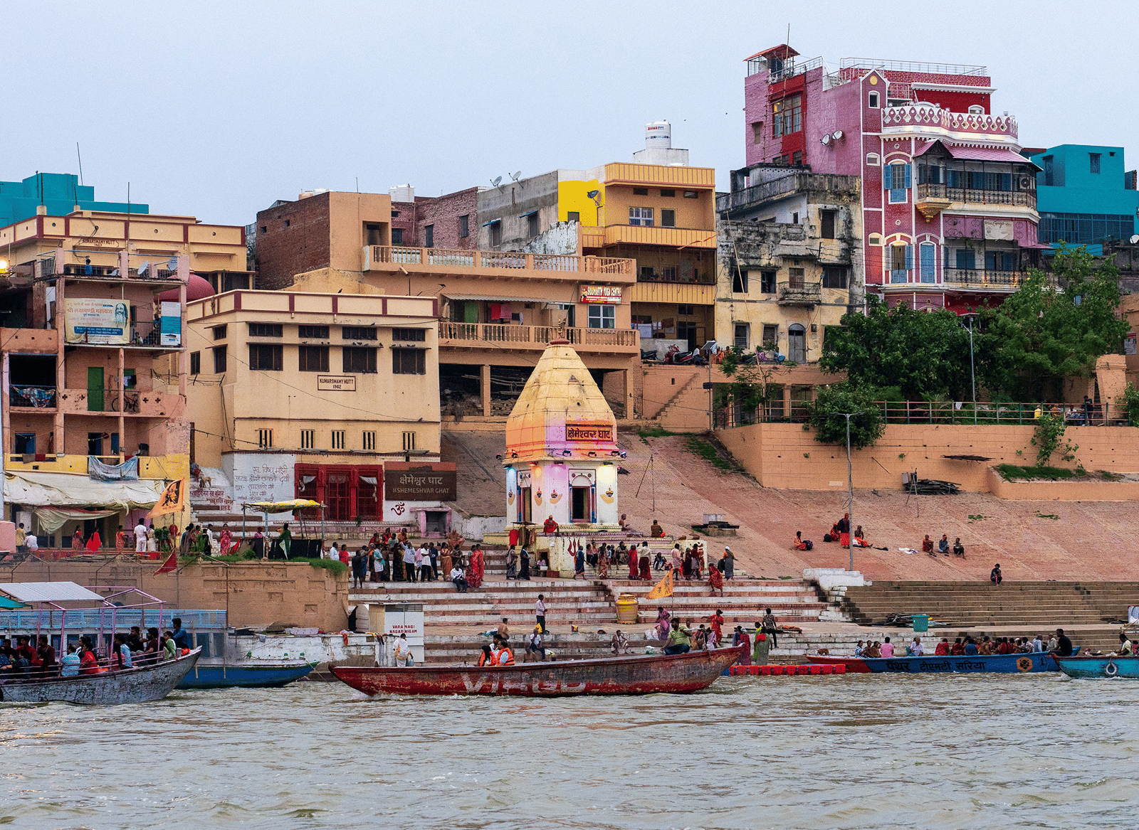 A ghat on the Ganges in Varanasi with a small domed shrine, stone steps, colourful riverside buildings, and wooden boats carrying visitors.