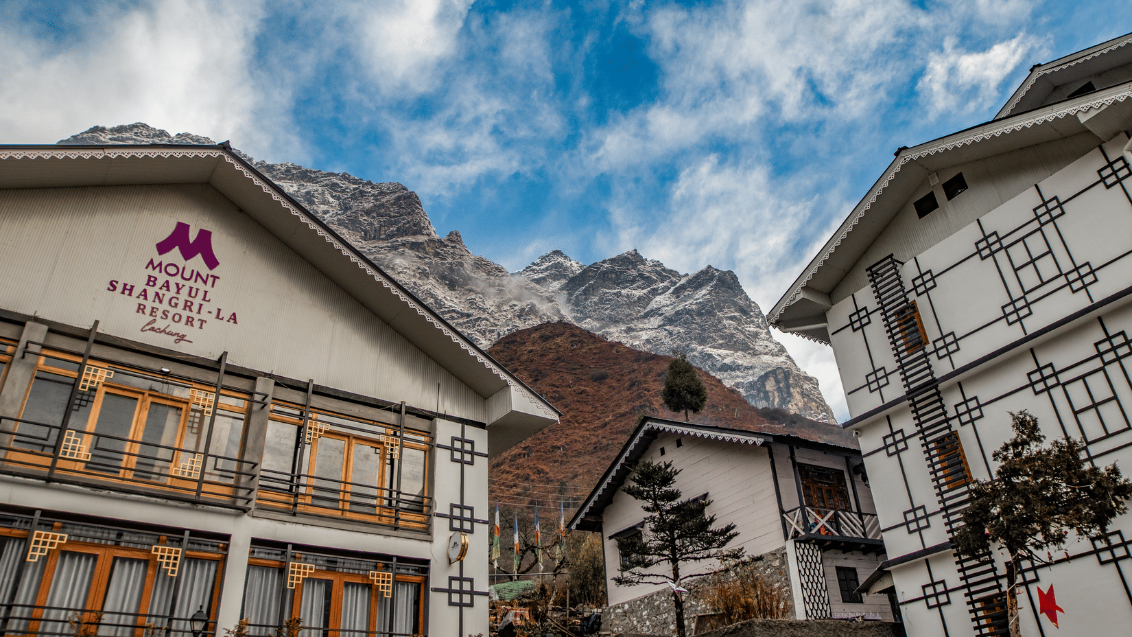 Mount Bayul Shangri-La Resort in Lachung features traditional-style buildings with wooden balconies, set against a mountainous backdrop beneath a partly cloudy sky.