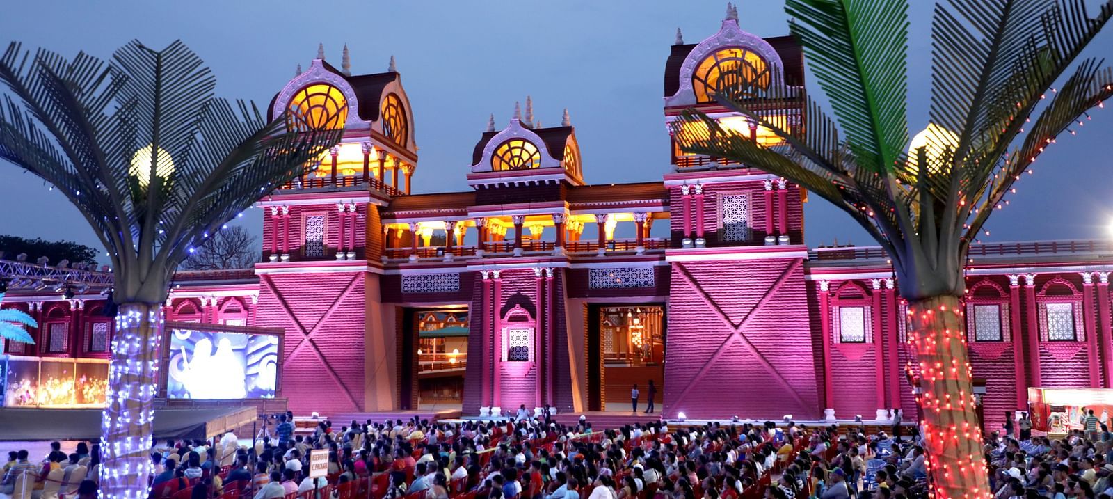 A large crowd gathers in front of a grand, illuminated building with arches and towers, surrounded by decorative palm trees. The structure is bathed in pink and purple lights, creating a festive atmosphere at Ramoji Film City.