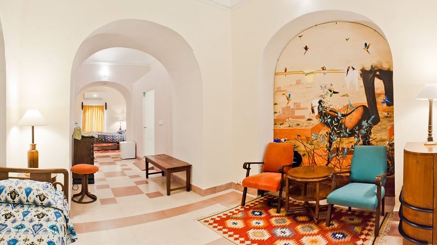 Hotel suite with arched doorways, a bedroom with a bed, a seating area with colorful chairs and artwork, and a hallway with benches at Tijara Fort-Palace, Alwar.