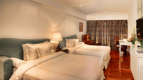 A club room with twin beds with a padded headboard, bedside lamps, a wooden floor, a writing desk with chair, a wall-mounted television, a mirror panel, and a large window with curtains at La Marvella.