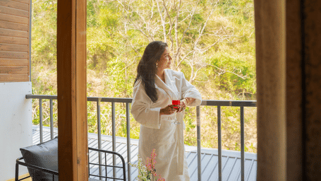 A person in a robe stands on a balcony at Perfect Stayz The Jungle Resort, observing the scenery.