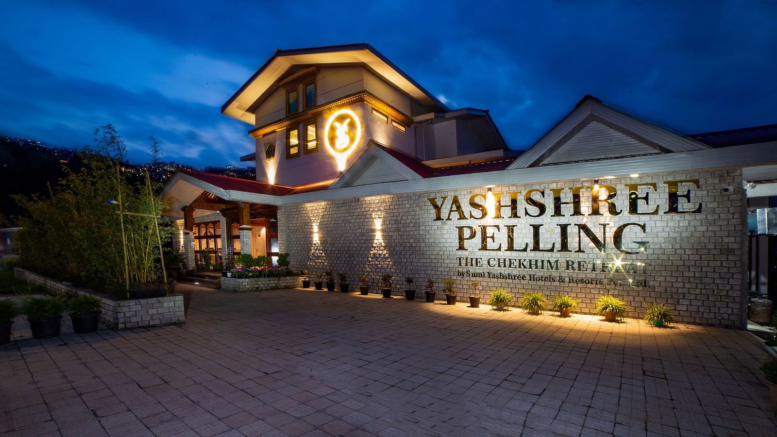 Evening view of Yashshree Pelling hotel, featuring a brightly lit entrance, stone façade, and red-roofed building under a deep blue sky.