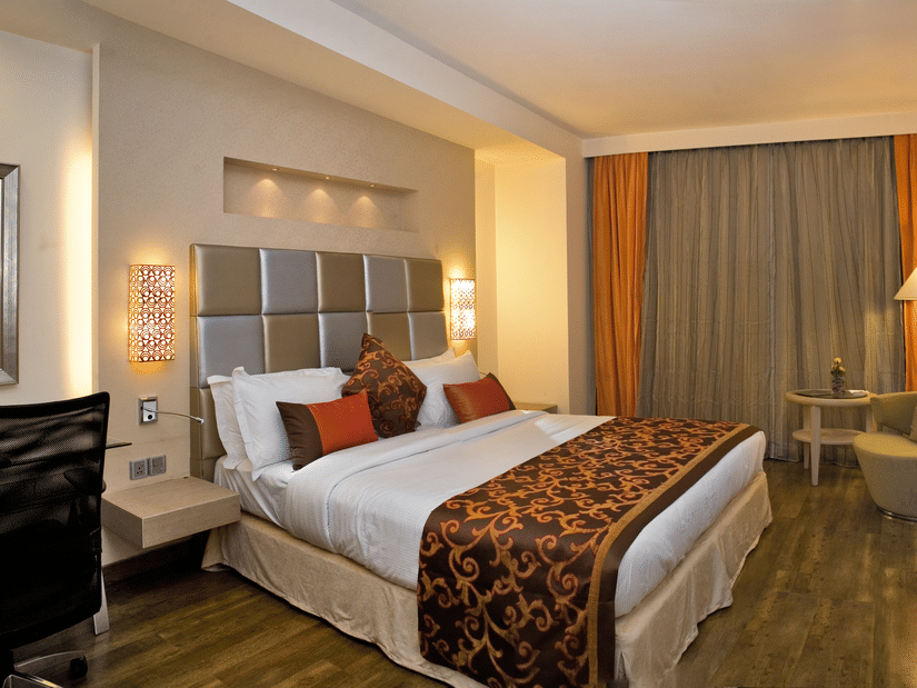 A room at The Sonnet, Kolkata featuring a bed with a patterned runner, a desk, a television, and large windows with curtains.