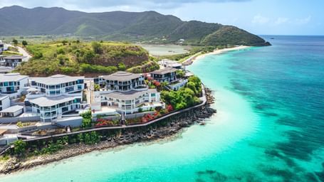 An aerial view of a Tamarind Hills Resort and Villas, Antigua with buildings perched on a rocky coastline, overlooking clear waters and surrounded by hills.