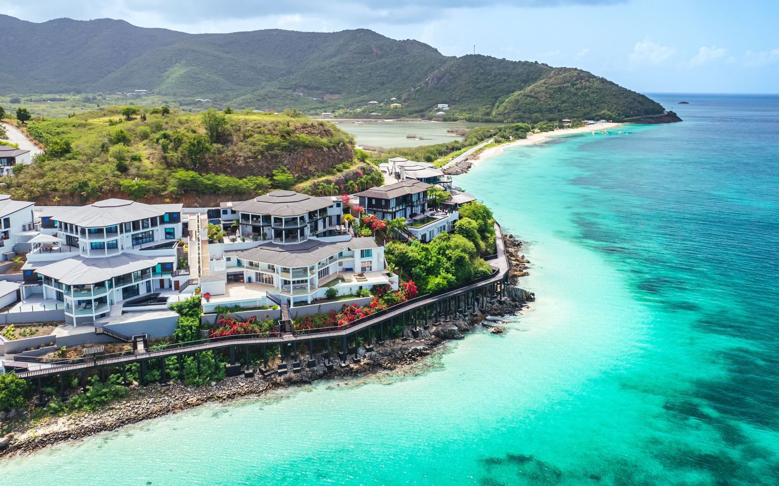 An aerial view of a luxury seaside resort with modern white buildings perched on a rocky coastline, overlooking turquoise waters and surrounded by lush green hills - Tamarind Hills Resort and Villas, Antigua.