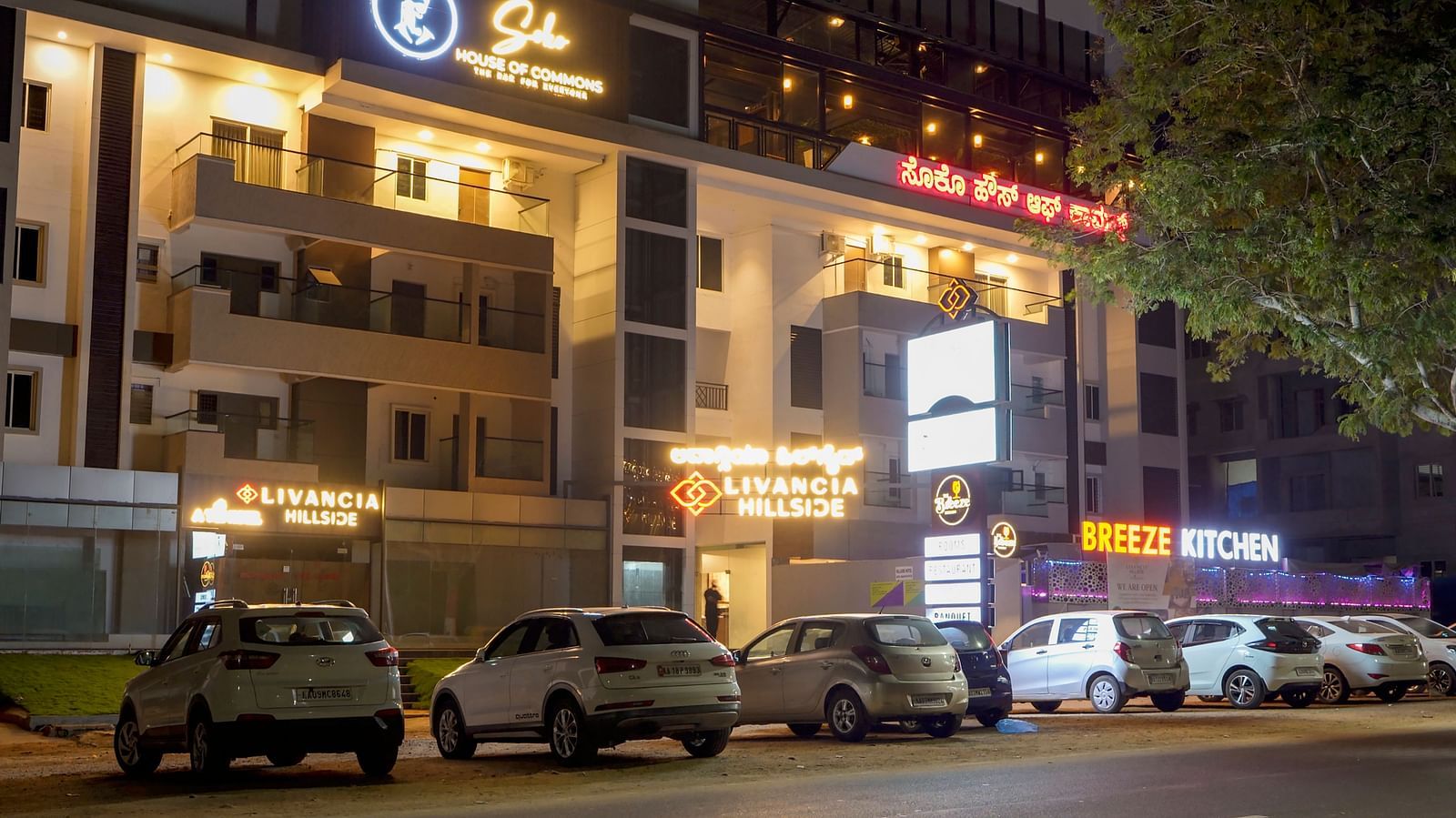 Night view of the hotel exterior with parked cars and lit-up signs along the street at TGI Livancia Hillside, JC Nagar, Mysore