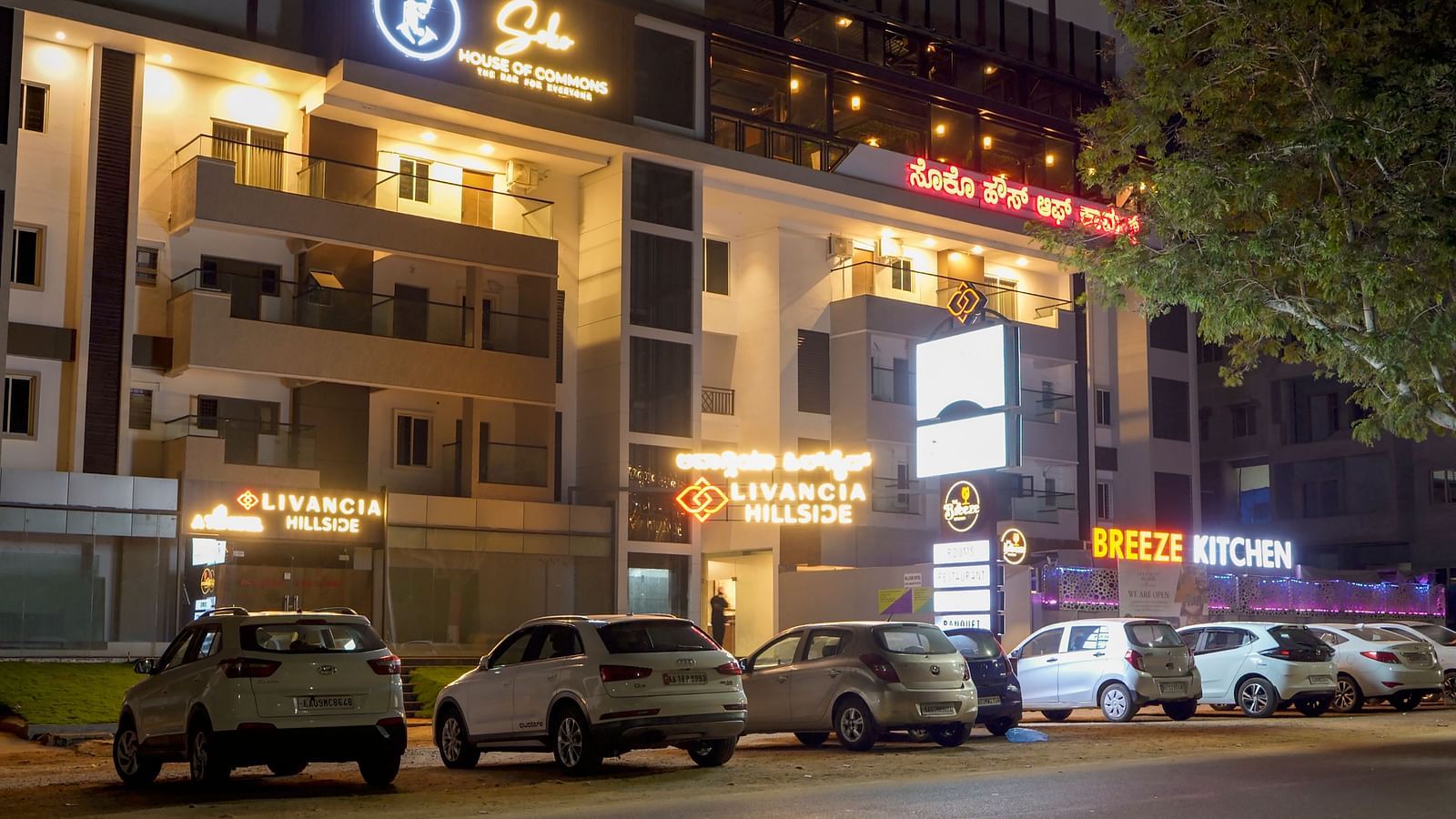 Night view of the hotel exterior with parked cars and lit-up signs along the street at TGI Livancia Hillside, JC Nagar, Mysore