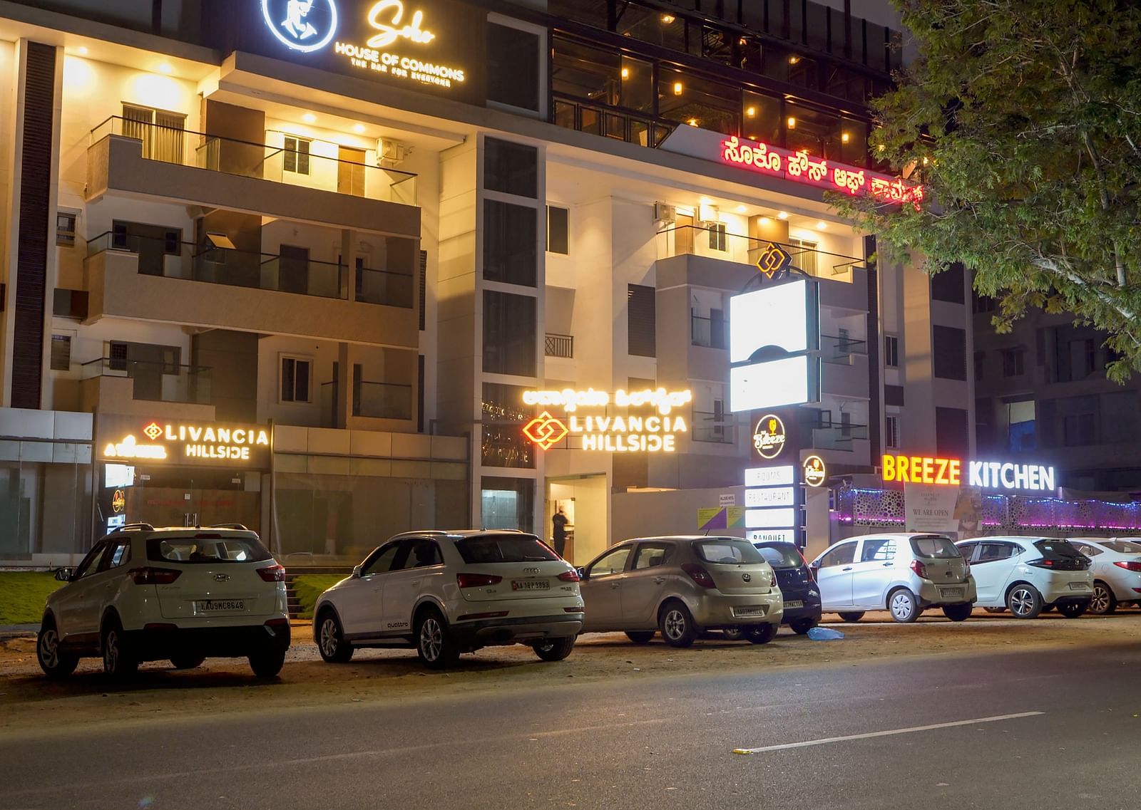 Night view of the hotel exterior with parked cars and lit-up signs along the street at TGI Livancia Hillside, JC Nagar, Mysore