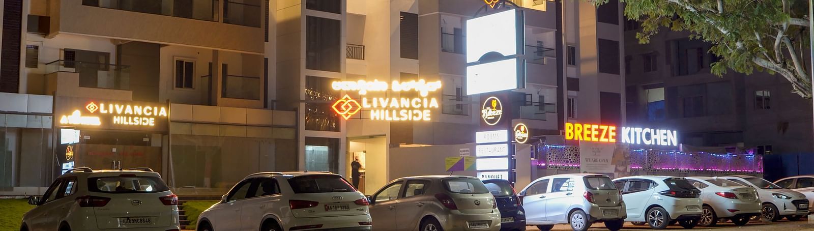 Night view of the hotel exterior with parked cars and lit-up signs along the street at TGI Livancia Hillside, JC Nagar, Mysore
