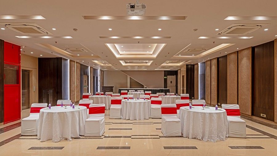 A conference hall with a projector screen at one end, multiple round tables surrounded by white-covered chairs, each with a red ribbon tied on the back.