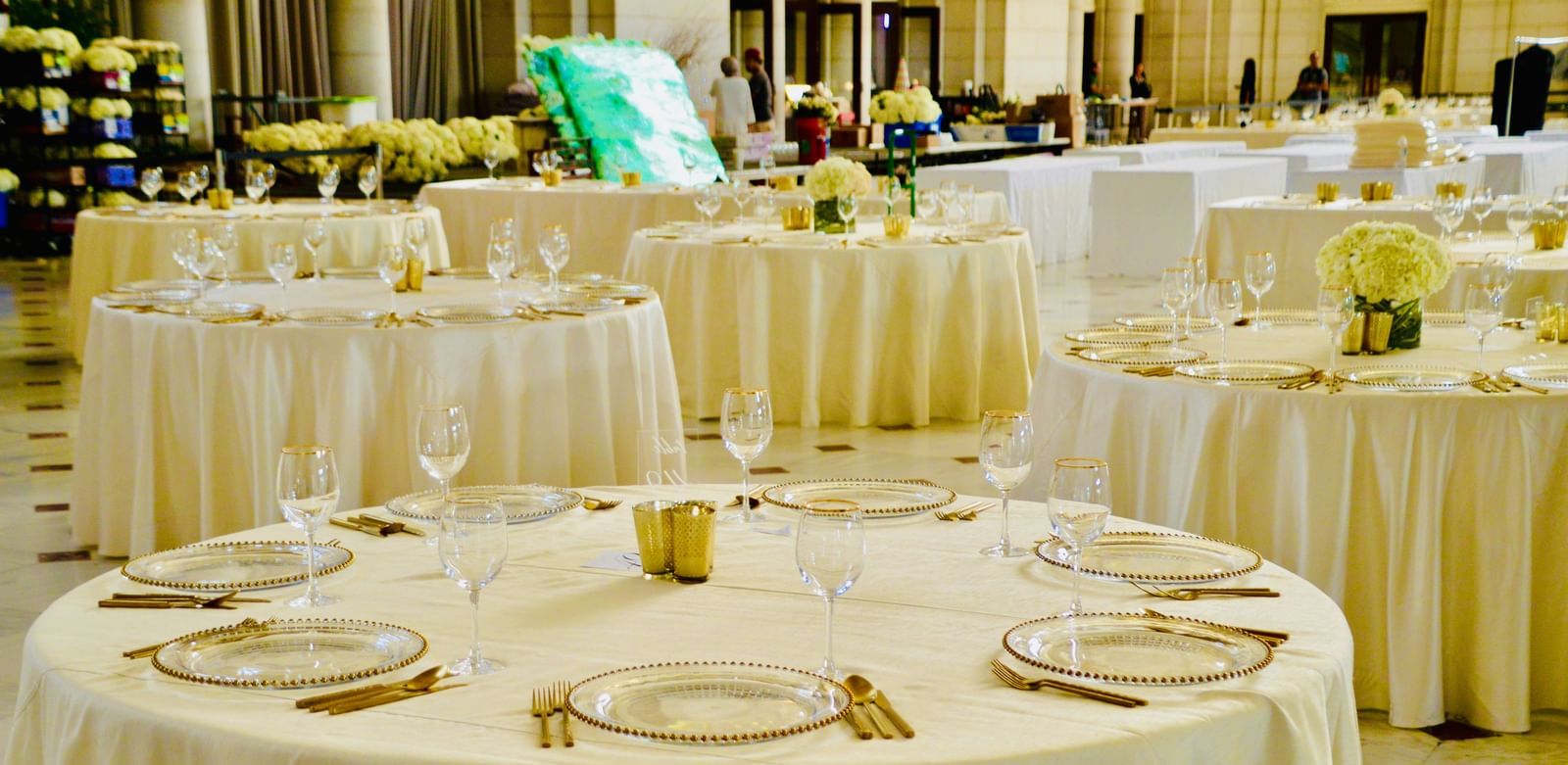 A banquet hall with round tables covered in white tablecloths, neatly arranged with plates, glasses, and cutlery for dining.