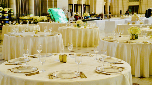 A beautifully arranged banquet hall with round tables covered in white linens and decorated with glassware.