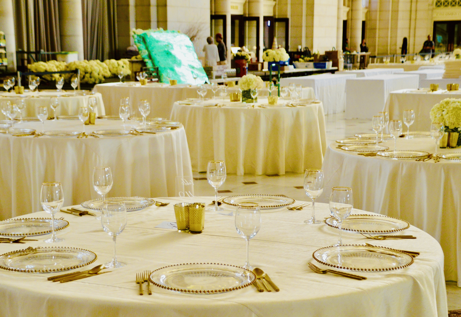 Close-up of a formally set dinner table for an event, featuring white tablecloths, gold-rimmed crockery, and a floral centrepiece.