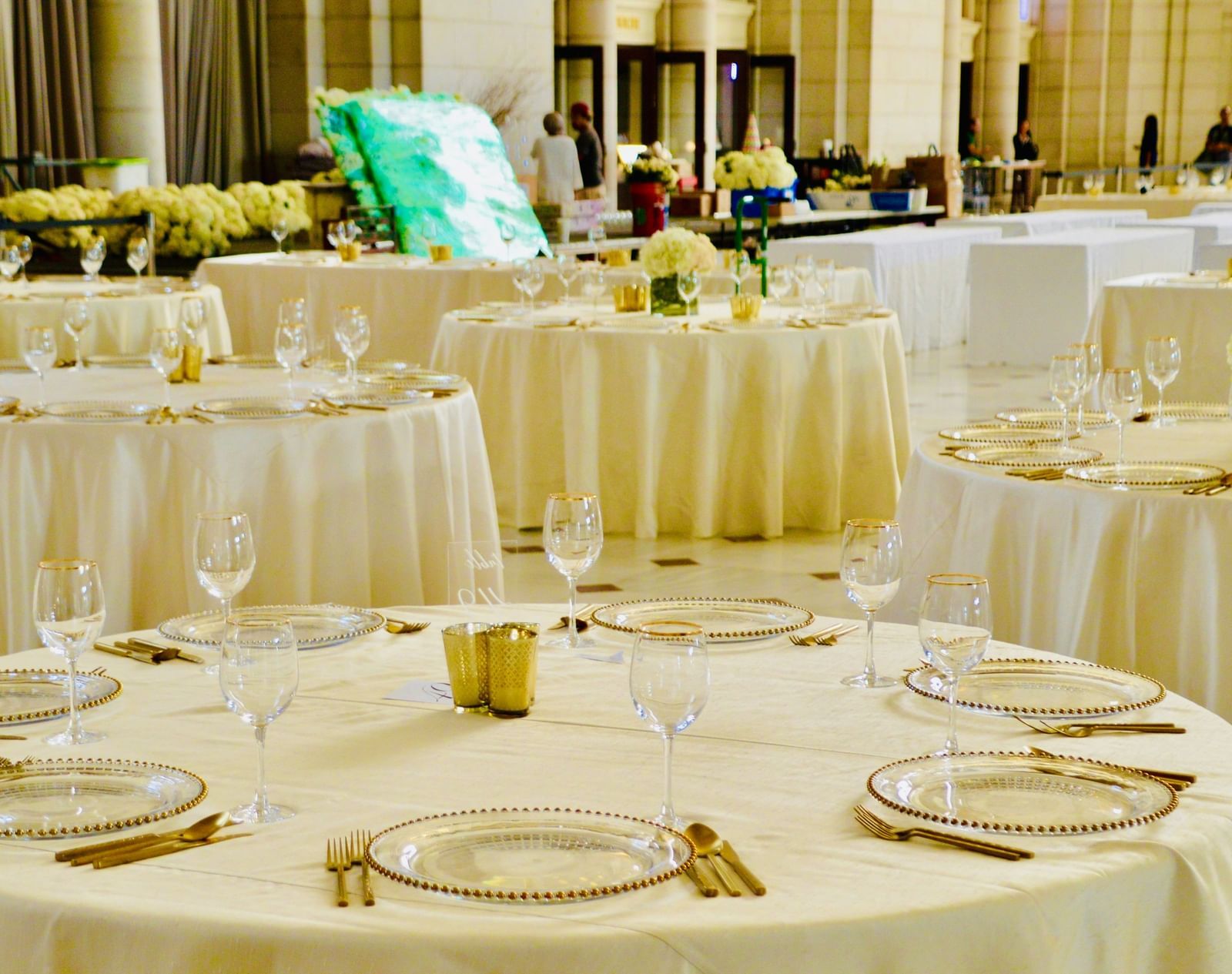 A formal dining setup showing multiple round tables draped in white cloth, set with silverware, gold-rimmed plates, and wine glasses, ready for an event.