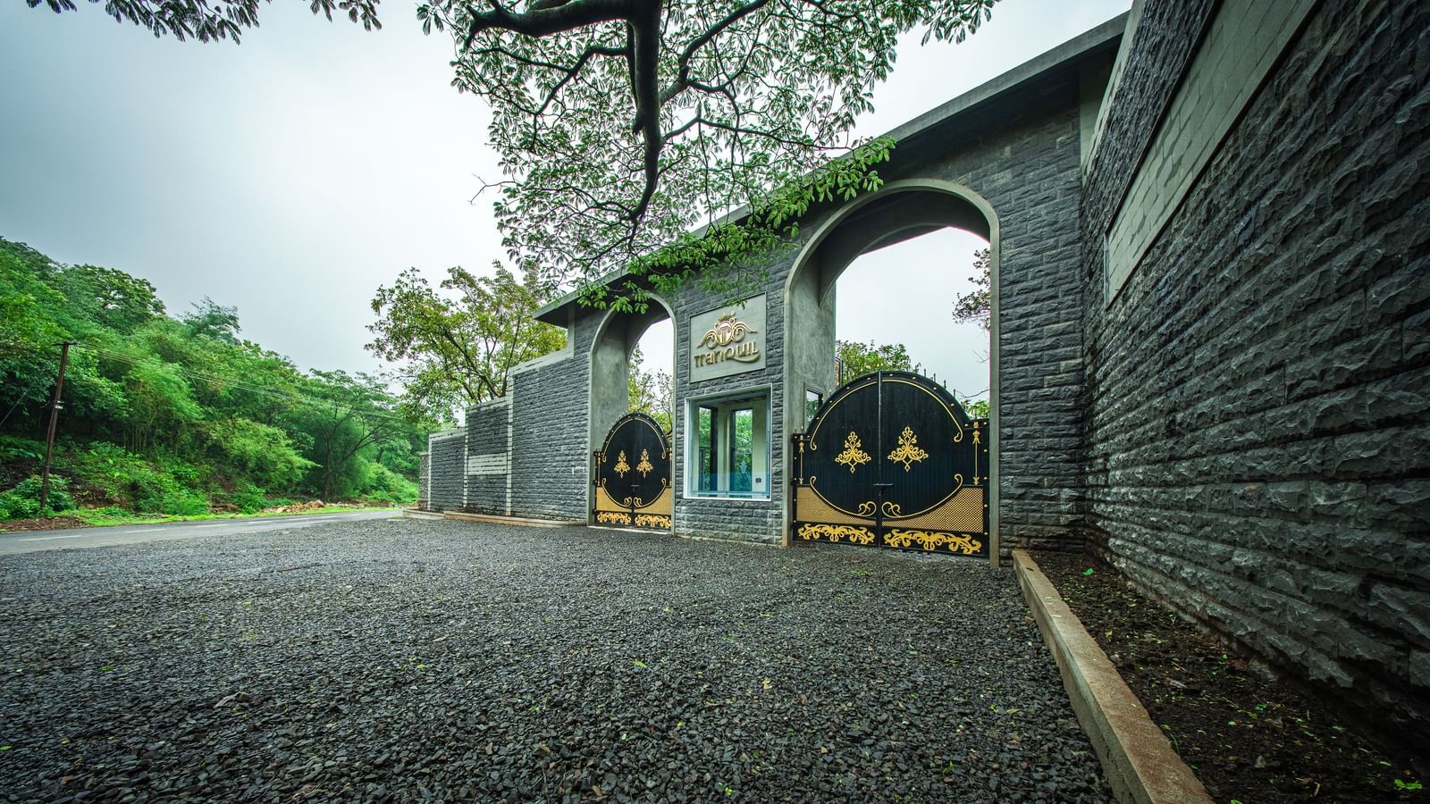 A stone gateway entrance with an arched top with lush greenery around - Tranquil Piedmont Estates & Resort, Lonavala