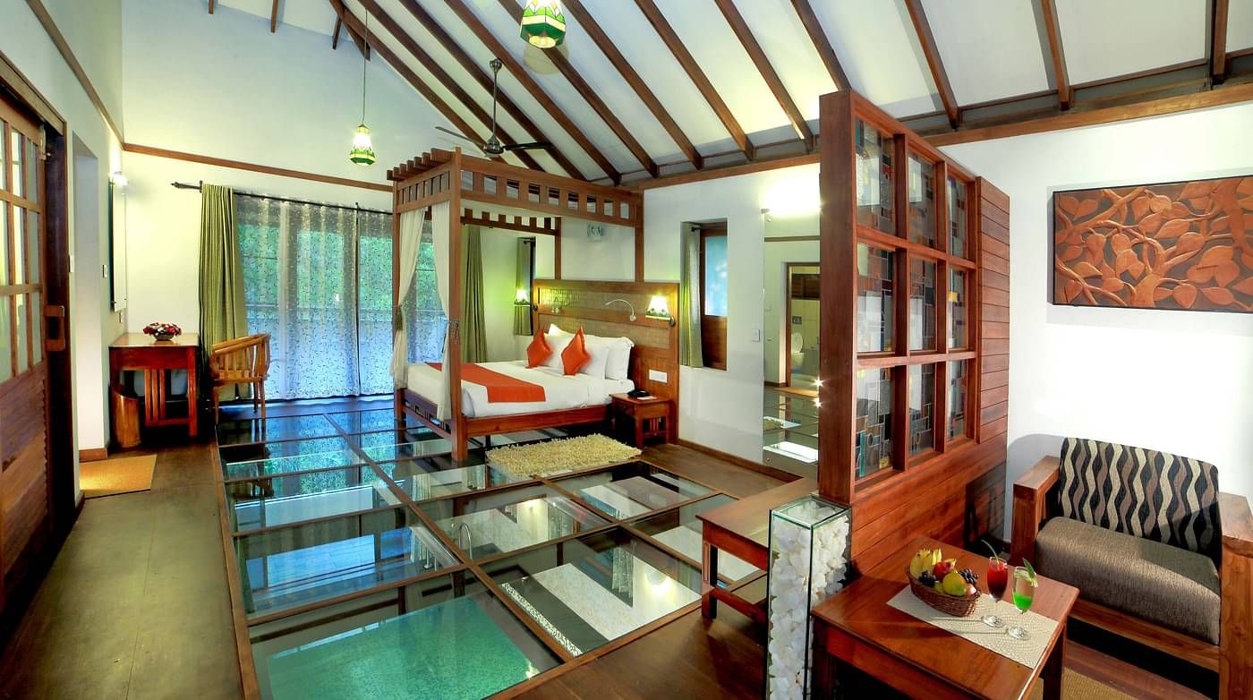 A full view of a glass-floored room that has a bed and other wooden furnishings | Vythiri Resort. Wayanad