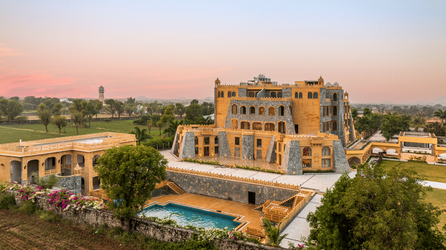 A high angle view of the facade of EsthereaRaj Leela, Ranakpur, featuring the outdoor swimming poo and surrounding green fields.
